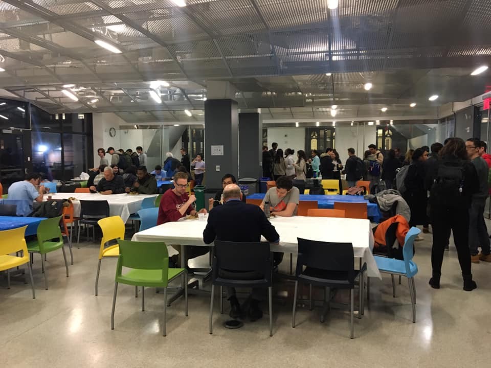 Student and professors standing and seated at various tables across a large room with pillars, tables and multi colored chairs all to meet informally at Got FU'd event.